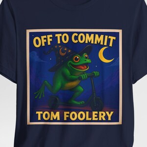 May include: Navy blue t-shirt with a green frog wearing a witch's hat and riding a scooter. The graphic reads "OFF TO COMMIT TOM FOOLERY" in yellow. A yellow crescent moon is in the background.