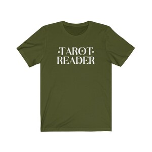 Op de afbeelding: Olijfgroen t-shirt met de witte tekst "TAROT READER". De tekst is gecentreerd en heeft steraccenten. Het shirt heeft korte mouwen en een ronde hals.