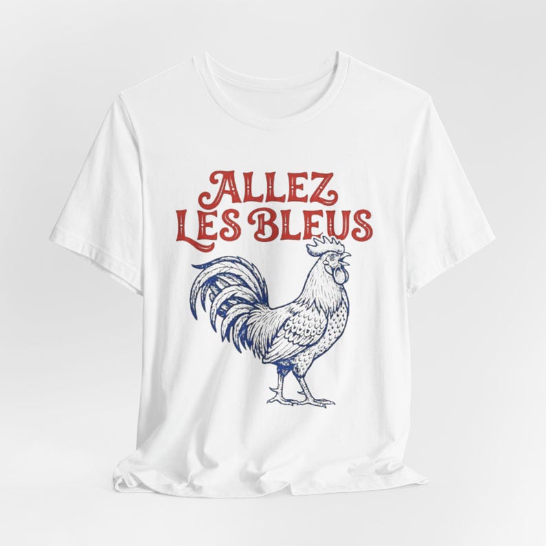 May include: A white short-sleeved t-shirt featuring a blue line-art illustration of a rooster and the red text "ALLEZ LES BLEUS" printed above the bird. The design is centered on the chest of the garment, which has a crew neckline and short sleeves.