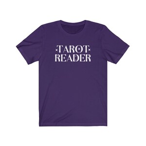 Op de afbeelding: Een paars t-shirt met de woorden "TAROT READER" in het wit gedrukt. De tekst is gecentreerd en heeft ster- en maanaccenten. Het shirt is gemaakt van een zacht materiaal.
