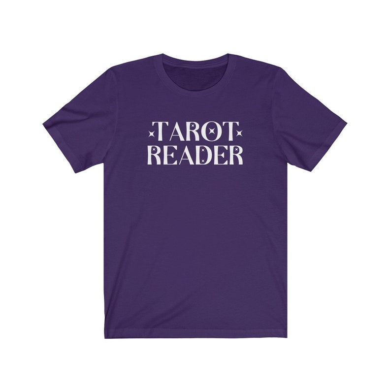 Op de afbeelding: Een paars t-shirt met de woorden "TAROT READER" in het wit gedrukt. De tekst is gecentreerd en heeft ster- en maanaccenten. Het shirt is gemaakt van een zacht materiaal.