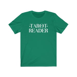 Op de afbeelding: Een groen t-shirt met de witte tekst "TAROT READER". De tekst is versierd met sterren en halve maan motieven. Het t-shirt is effen van kleur en lijkt gemaakt van een zacht materiaal.