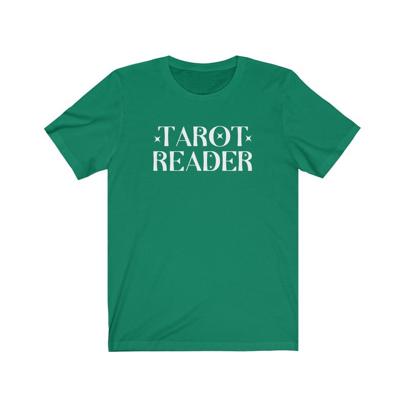 Op de afbeelding: Een groen t-shirt met de witte tekst "TAROT READER". De tekst is versierd met sterren en halve maan motieven. Het t-shirt is effen van kleur en lijkt gemaakt van een zacht materiaal.