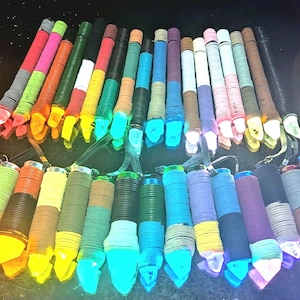 Könnte beinhalten: Eine Sammlung farbenfroher, zylindrischer LED-Leuchten mit verschiedenfarbigen Wicklungen. Jede Leuchte hat ein klares, spitzes Ende und eine kleine Schlaufe zum Aufhängen. Die Leuchten sind auf einer dunklen Oberfläche angeordnet, einige leuchten hell.