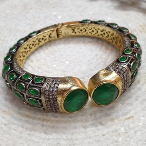 May include: A gold and silver hinged bracelet with emerald green stones. The bracelet has a gold interior with intricate cut-out designs. The ends feature large, oval green stones. The text "BA067/JJI-085 all size available openable" is visible.