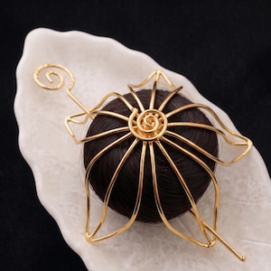 May include: A gold-coloured hair accessory with a floral design, featuring a spiral centre and radiating lines. The accessory is set against a dark brown hair bun on a white, leaf-shaped dish, with a black background.