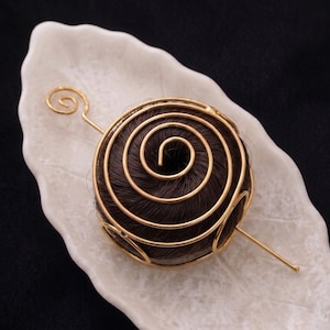Könnte beinhalten: Ein einzigartiges Haaraccessoire mit einem dunkelbraunen, kreisförmigen Design und einem Spiralmuster, das von einem goldfarbenen Drahtrahmen umschlossen ist. Das Haarteil liegt auf einer weißen, blattförmigen Schale vor schwarzem Hintergrund.