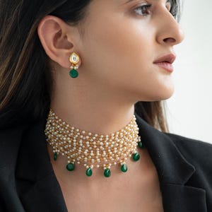 Può includere: Una collana girocollo dorata con più fili di piccole perle bianche e gemme a goccia verde smeraldo. Sono visibili anche orecchini dorati abbinati con gemme a goccia verde smeraldo.