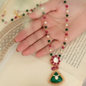 May include: A gold necklace with a pendant featuring a green triangular stone, surrounded by small white stones. The necklace has a chain of red, green, and white beads. The pendant is held by a hand.