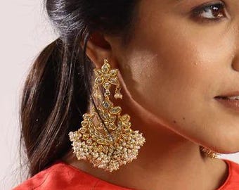 May include: Gold-toned chandelier earrings with intricate filigree and pearl accents. The earrings are worn by a person wearing a red top. The earrings have a detailed design and a cluster of small pearls at the bottom.