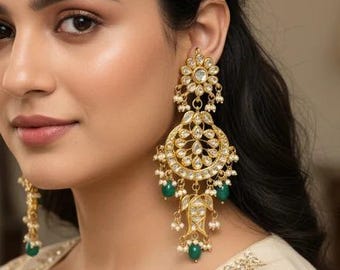 May include: A pair of gold-toned chandelier earrings featuring clear, teardrop-shaped stones arranged in a circular pattern, accented with small white pearls and dangling green beads, worn with a cream-colored garment with gold embroidery.