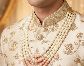 Puede incluir: Un sherwani de color crema con intrincados bordados florales dorados y cuello alto, combinado con un collar de varias hileras compuesto por perlas blancas ovaladas y cuentas redondas más pequeñas de color melocotón sobre el pecho.