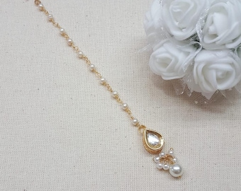 May include: A gold-colored headpiece with a teardrop-shaped pendant and a chain of small pearls. The pendant has a clear, faceted stone. A cluster of pearls dangles below the pendant. The headpiece is designed to be worn on the forehead.