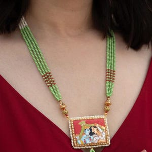 May include: A multi-strand necklace featuring light green beads with small red accents, connected to a rectangular pendant. The pendant displays a painted scene of two figures in traditional attire on a red background, framed by tiny white beads.