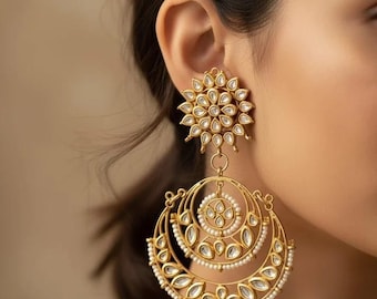 May include: A large gold-toned earring featuring a floral-shaped stud with white stone inlays. A circular pendant hangs below, decorated with concentric rings, teardrop-shaped white stones, and small white beads along the edges.