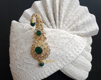 Puede incluir: Turbante blanco con un alfiler de oro y verde adornado con piedras preciosas. El alfiler tiene tres piedras verdes y está decorado con diamantes de imitación transparentes. El turbante está hecho de una tela texturizada.