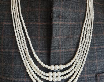 Op de afbeelding: Een meerrijige parelketting met een groot parelmiddelpunt. De ketting is gemaakt van witte parels en wordt gedragen op een grijs geruit overhemd.