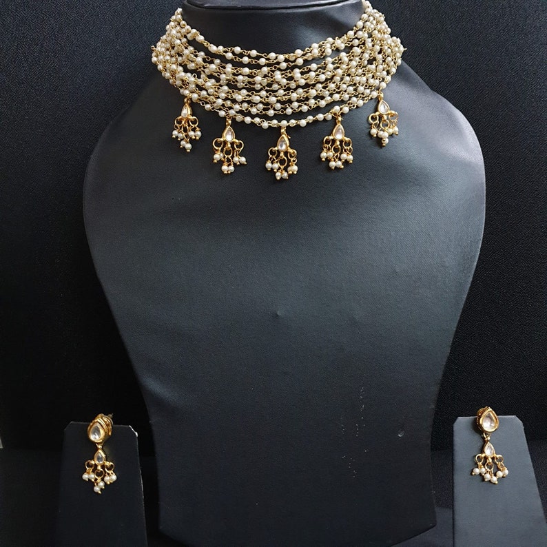 May include: A gold choker necklace with multiple strands of white pearls and dangling pearl and stone accents. Matching earrings with pearl and stone accents.