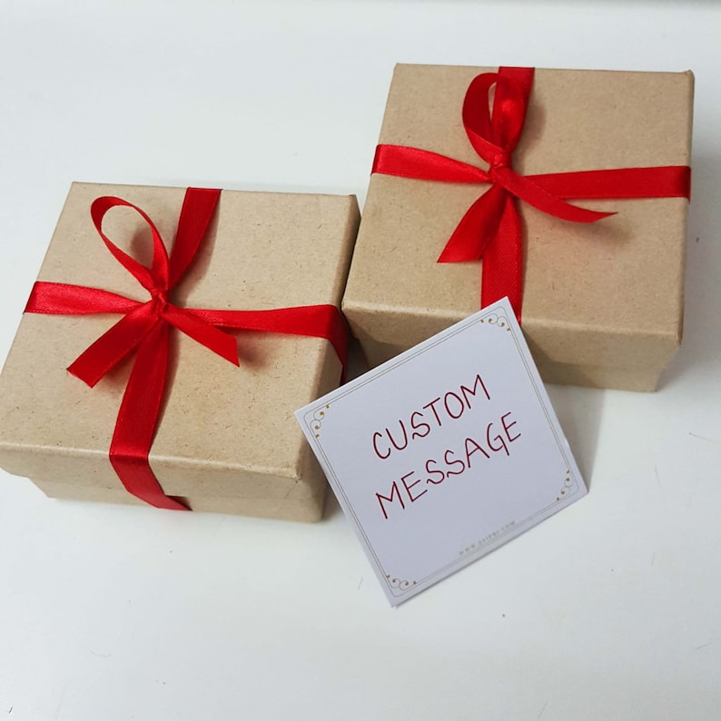 Pu&ograve; includere: Due piccole scatole regalo marroni legate con fiocchi di nastro rosso. Un biglietto bianco con il testo "MESSAGGIO PERSONALIZZATO" &egrave; davanti alle scatole.