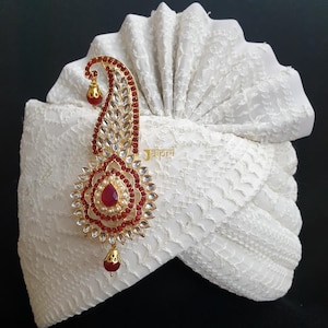 May include: A white turban with intricate lace-like detailing, adorned with a gold-toned, ornate brooch. The brooch features red and clear gemstones, and a central teardrop-shaped red stone. The word "jaipri" is visible on the turban.