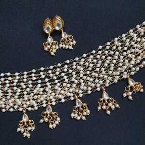 May include: A gold necklace with multiple strands of small white pearls and teardrop-shaped pendants with clear stones. The necklace has a matching pair of earrings with the same design.