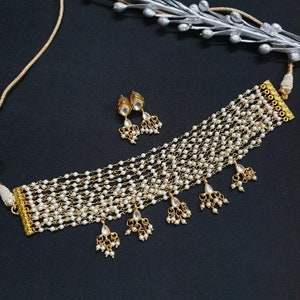 May include: A gold choker necklace with multiple strands of white pearls and dangling pearl accents. The necklace comes with a matching pair of earrings with pearl accents.