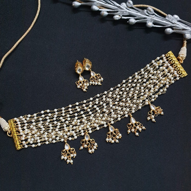 May include: A gold choker necklace with multiple strands of white pearls and dangling pearl accents. The necklace comes with a matching pair of earrings with pearl accents.