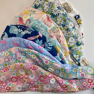 May include: A stack of 10 colorful fabric scrub hats with various patterns, including flamingos, pineapples, flowers, and a cartoon character. The hats have a button on the side and the text "Poor Sweet" is visible on the bottom hat.