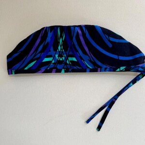 May include: A patterned surgical scrub cap in shades of blue, teal, and purple on a black background. The cap has a tie closure. The fabric features an abstract design of curved lines and geometric shapes.