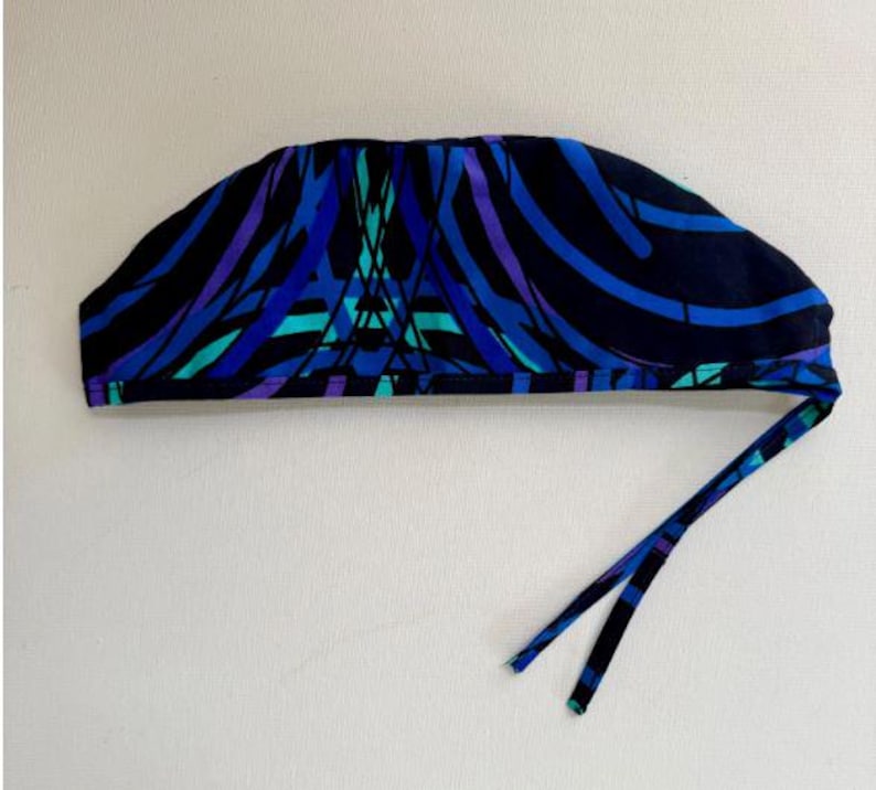 May include: A patterned surgical scrub cap in shades of blue, teal, and purple on a black background. The cap has a tie closure. The fabric features an abstract design of curved lines and geometric shapes.