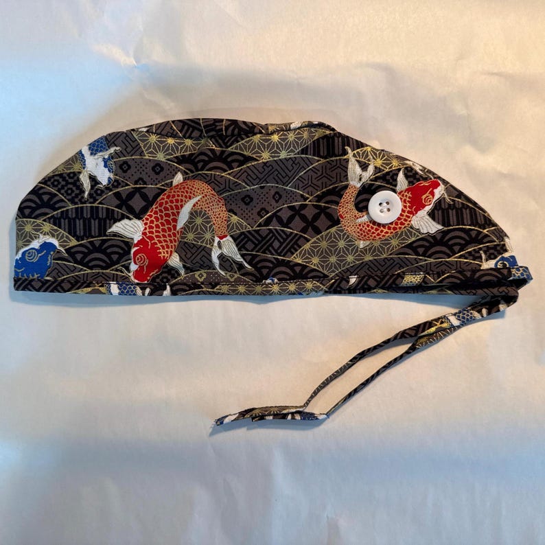 May include: A patterned surgical scrub cap featuring a koi fish design. The cap is primarily black with gold, white, and red accents. It has a white button and tie-back straps for an adjustable fit. The koi fish are swimming across a wave pattern.