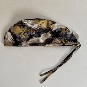 May include: A black surgical cap with a Japanese crane and floral pattern. The cap features white cranes with outstretched wings, gold and gray chrysanthemums, and a tie closure. The fabric has a repeating design.