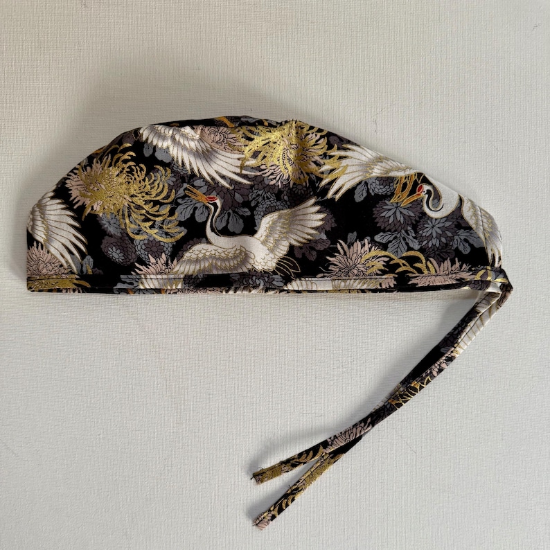 May include: A black surgical cap with a Japanese crane and floral pattern. The cap features white cranes with outstretched wings, gold and gray chrysanthemums, and a tie closure. The fabric has a repeating design.