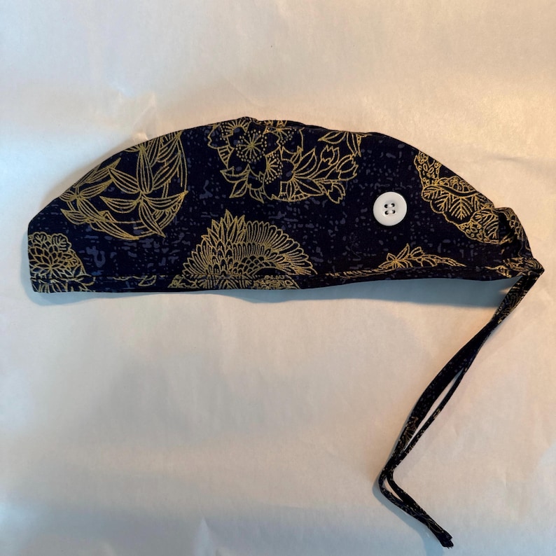 May include: A navy blue surgical scrub cap with a gold floral pattern and a white button. The cap has a tie in the back for an adjustable fit. The fabric has a subtle, textured appearance.