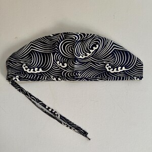 May include: A navy blue and white patterned cap with a wave design. The cap has a tie closure and is made of fabric. The wave pattern is a repeating design of curved lines.