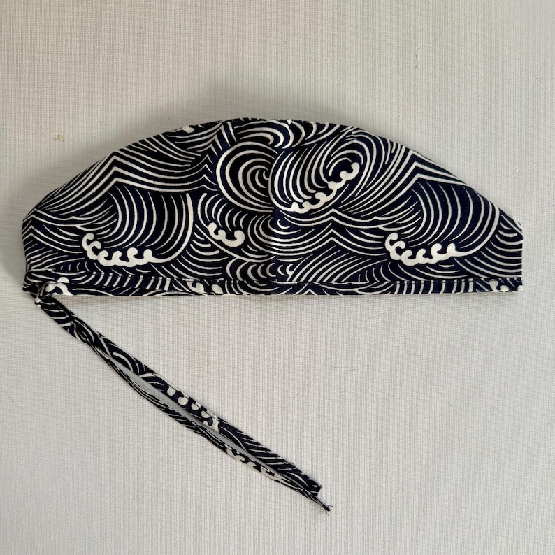 May include: A navy blue and white patterned cap with a wave design. The cap has a tie closure and is made of fabric. The wave pattern is a repeating design of curved lines.