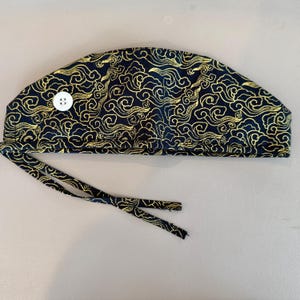 May include: A navy blue surgical scrub cap with a gold cloud pattern. The cap has a white button and tie strings. The fabric is patterned with swirling gold clouds.