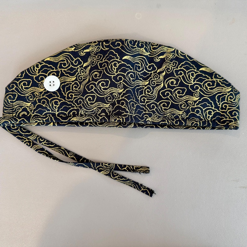 May include: A navy blue surgical scrub cap with a gold cloud pattern. The cap has a white button and tie strings. The fabric is patterned with swirling gold clouds.
