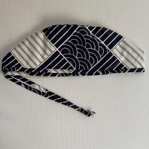 May include: A navy blue and white patterned surgical scrub cap. The cap features a geometric design with stripes, squares, and semi-circles. The cap has a long tie for securing it in place.