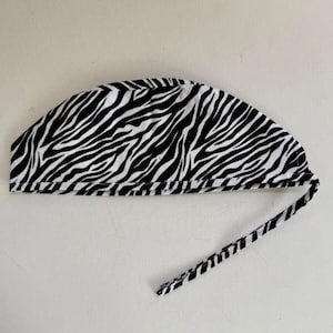 May include: A black and white zebra print surgical cap with a matching tie. The cap is made of fabric and is designed to cover the head. The zebra pattern consists of black stripes on a white background.