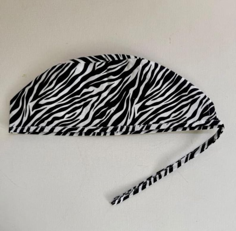 May include: A black and white zebra print surgical cap with a matching tie. The cap is made of fabric and is designed to cover the head. The zebra pattern consists of black stripes on a white background.