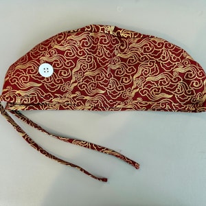 May include: A red surgical scrub cap with a gold swirling cloud pattern. The cap has a white button and matching red ties. The fabric is a rich red with gold accents.