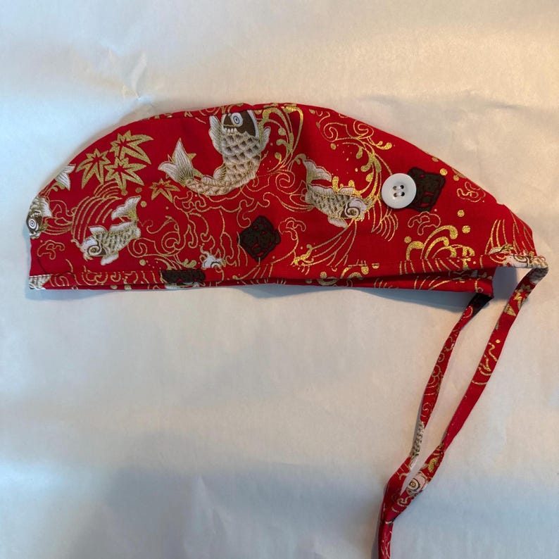 May include: Red surgical cap with a Japanese koi fish pattern. The cap features a repeating design of gold koi fish, maple leaves, and swirling patterns on a red background. It has a white button and red ties.