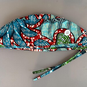 May include: A patterned surgical scrub cap with a white button and tie strings. The cap features a vibrant design with blue, red, and green abstract shapes on a light blue background. The ties are green and blue.