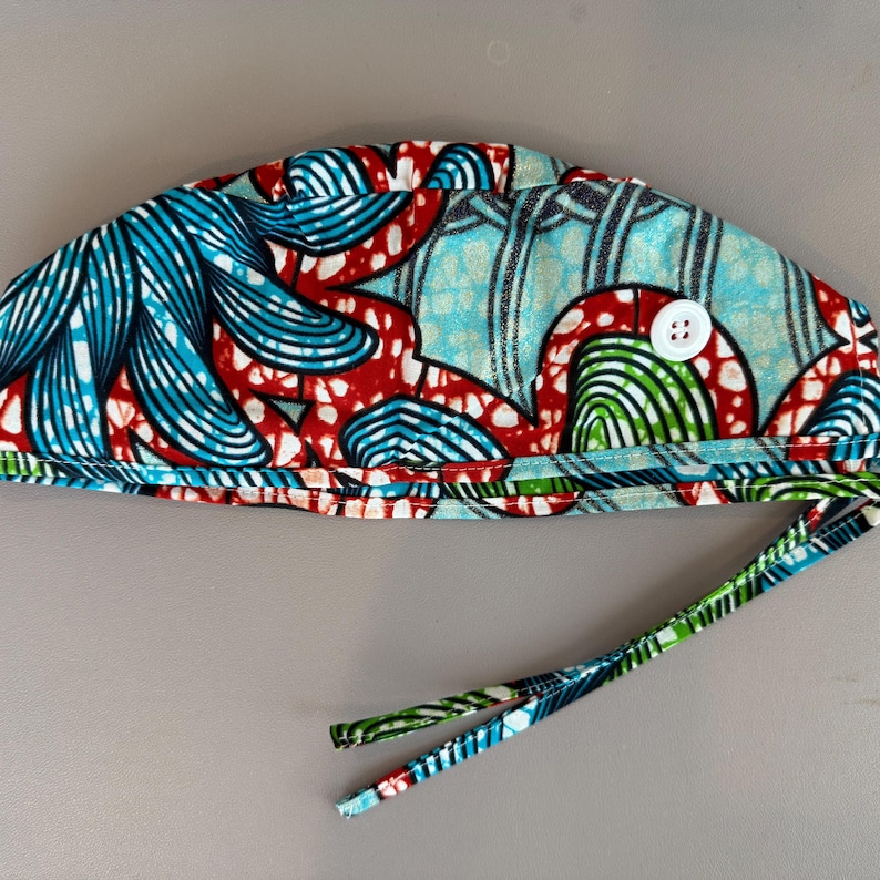 May include: A patterned surgical scrub cap with a white button and tie strings. The cap features a vibrant design with blue, red, and green abstract shapes on a light blue background. The ties are green and blue.