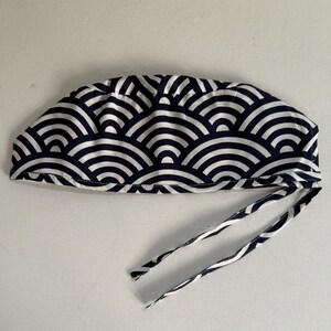 May include: A navy blue and white patterned surgical scrub cap with ties. The cap features a repeating wave-like design. The ties are made of the same fabric as the cap. The cap is designed to be worn in a medical setting.