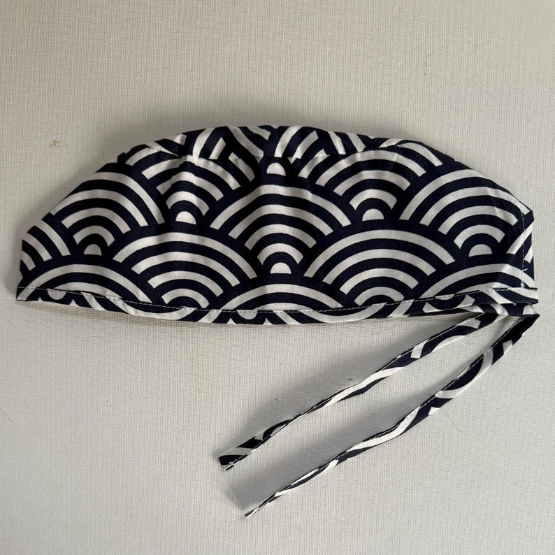 May include: A navy blue and white patterned surgical scrub cap with ties. The cap features a repeating wave-like design. The ties are made of the same fabric as the cap. The cap is designed to be worn in a medical setting.