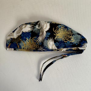 May include: A patterned surgical cap featuring a crane and floral design. The cap is primarily black with white, gold, and blue accents. It has a tie closure at the back for an adjustable fit.