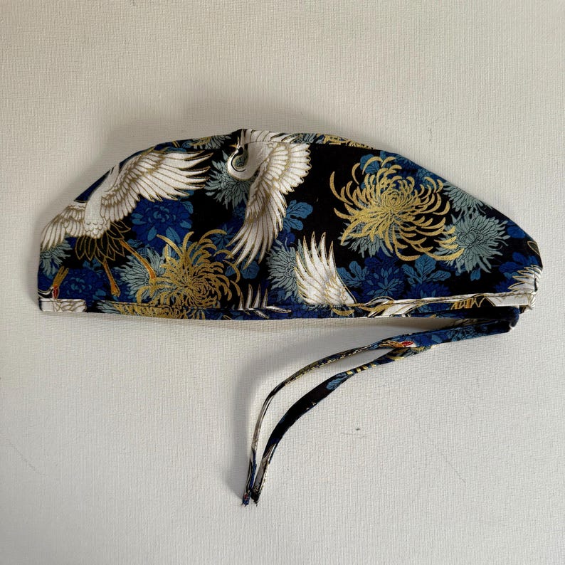 May include: A patterned surgical cap featuring a crane and floral design. The cap is primarily black with white, gold, and blue accents. It has a tie closure at the back for an adjustable fit.