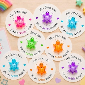 May include: Seven circular white cards each featuring a small translucent plastic turtle in pink, purple, blue, green, or orange. Each card displays the text Mrs Jones says you are turtely awesome with small heart icons and radiating lines.